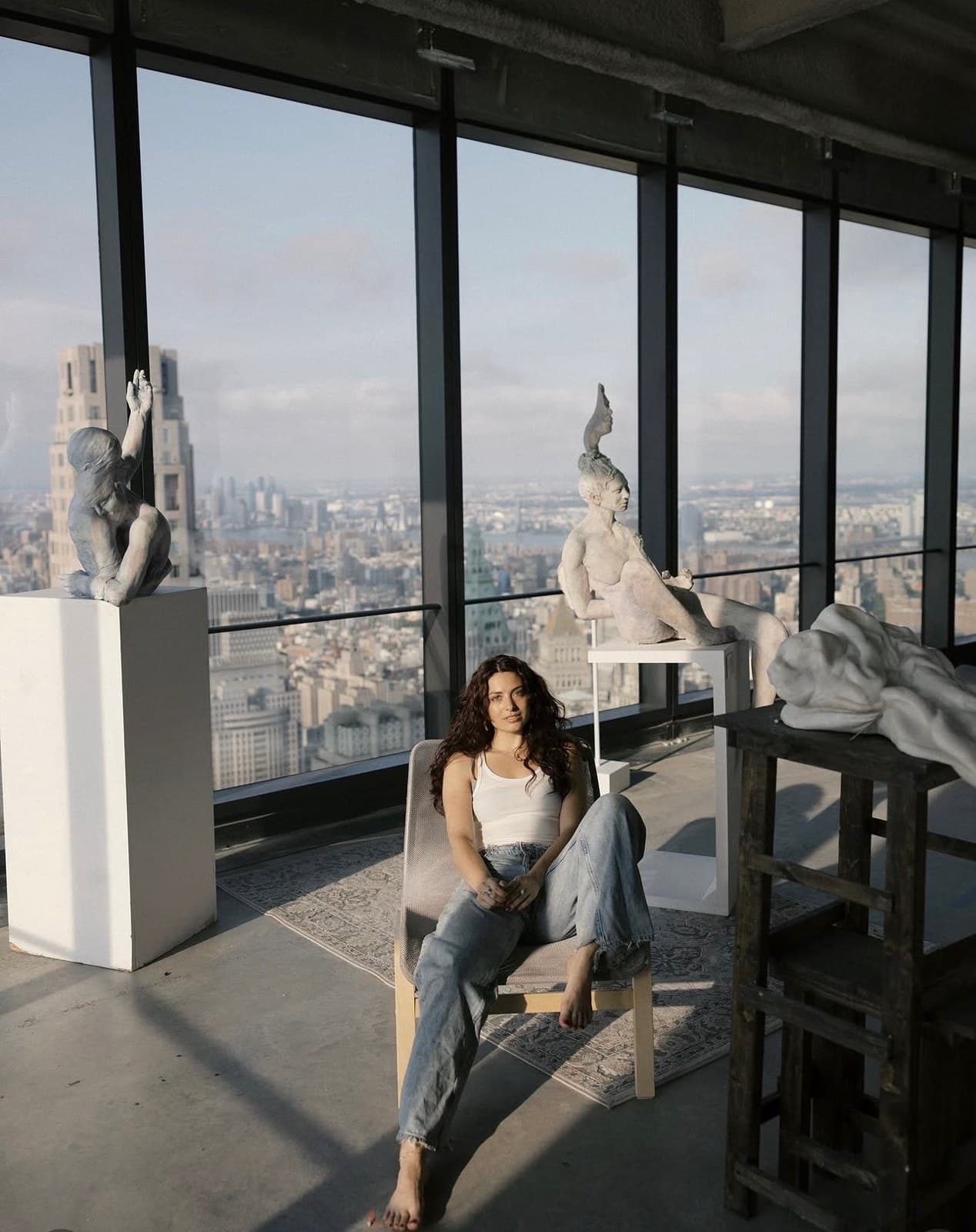 Artist Michela Roman seated among her sculptures in a studio overlooking the New York skyline