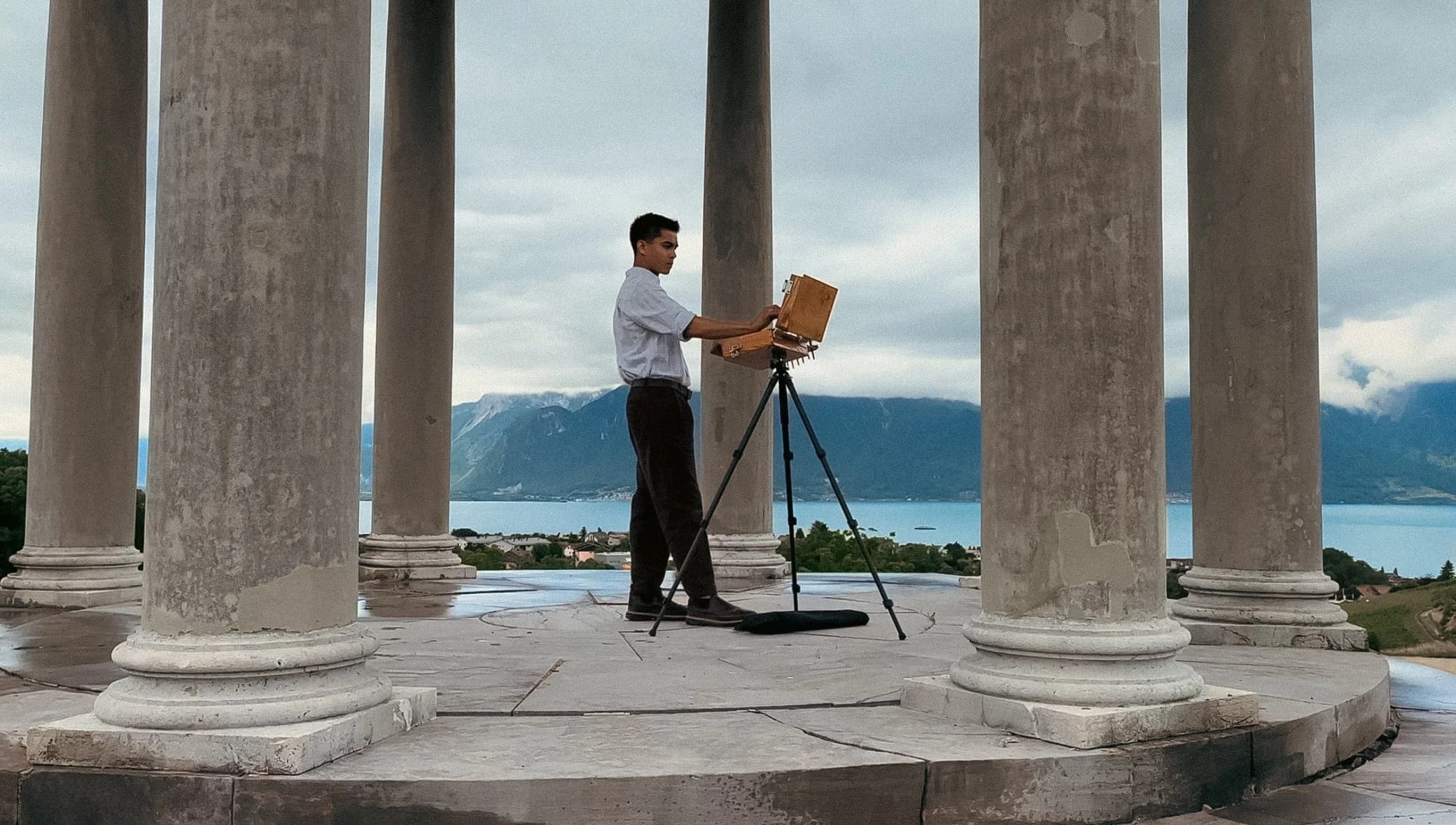Zachary Le painting at an easel between tall columns overlooking a lake and mountains